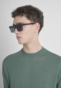 Suéter verde de punto con cuello redondo, que presenta un sutil logo. El modelo lleva gafas de sol rectangulares oscuras. Fondo claro.