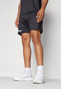 Zwarte sportshorts van lichtgewicht stof, met een gladde textuur en een wit logo aan de zijkant, gecombineerd met witte sportschoenen en sokken.