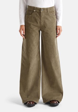 Person wearing wide-leg olive green corduroy pants, white long-sleeve top, and brown chunky shoes, standing against a plain background.