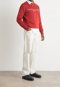 Czerwony sweter z logo "TOMMY HILFIGER", pod spodem biały służbowy koszula, białe spodnie i czarne obuwie; zaprezentowany strój casualowy.