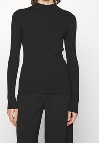 Pull en tricot noir à côtes avec un col montant, une silhouette ajustée et des manches longues, présentant des motifs texturés subtils sur les épaules.