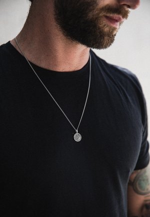 Sølvvedhængs halskæde på en sølvkæde, prydet med en rund skive med et detaljeret design, båret over en sort t-shirt.