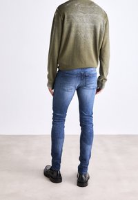Suéter verde oliva con una textura suave, combinado con jeans azules elásticos con un lavado desteñido y bolsillos traseros, usados con zapatos negros.