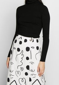 Kvinna som bär en svart långärmad polo och en vit kjol med abstrakt svart linjekonst och prickmönster.