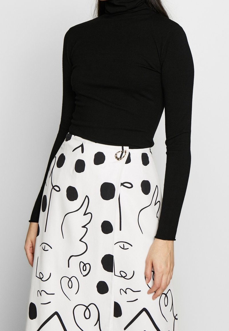 Kvinna som bär en svart långärmad polo och en vit kjol med abstrakt svart linjekonst och prickmönster.