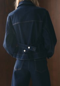 Giacca in denim scuro con design aderente, dettagli con bottoni e vita con cintura. Presenta cuciture sul giogo posteriore e un colletto classico.