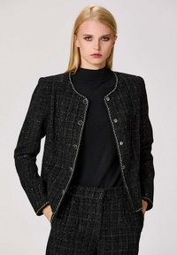 Veste en tweed noir avec un col rond, des boutons en argent et des détails de surfilage blancs contrastants, associée à un col roulé noir.