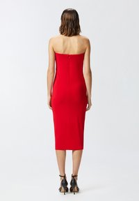 Crvena, bez naramenica, uska haljina do koljena s zatvaračem na leđima. Glatka tekstura, oblikovani dizajn. Crne štikle su vidljive.