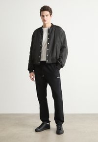 Svart bomberjakke med trykknapper, hvit t-skjorte, svarte sweatpants med logo, og svarte sneakers. Glatt stoff, minimalistisk design.
