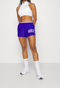 Bílý sportovní crop top se zipem, v kombinaci s fialovými šortkami Nike s logem. Oblečeno s bílými teniskami Nike a ponožkami do poloviny lýtek.