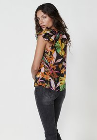 Blusa floreale con maniche a volant, caratterizzata da foglie e fiori colorati su uno sfondo nero. Abbinata a jeans in denim grigio scuro.