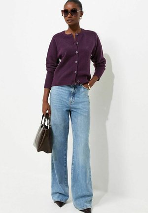 Femme portant de grandes lunettes de soleil, un cardigan violet boutonné, un jean bleu à jambes larges, tenant un sac à main marron, debout devant un fond blanc.