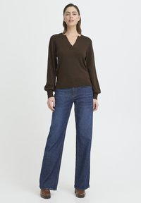 Smeđa džemper s V-izrezom i dugim, napuhanim rukavima, uparena s visokim strukom i širokim tamnoplavim trapericama. Nose se smeđe gležnjače.