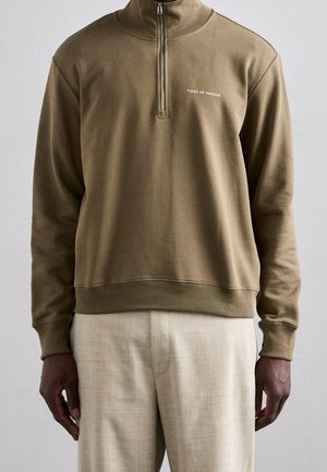 Man draagt een olijfgroene kwart-ritssweatshirt met het "Tiger of Sweden"-logo en beige linnen broek tegen een effen achtergrond.