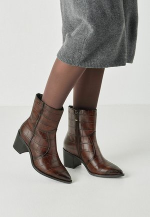 Bottines en cuir marron à texture crocodile avec talons carrés, portées avec des collants marron transparents et une jupe en tricot gris.