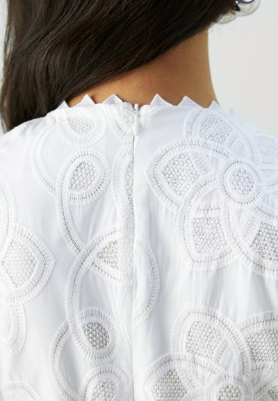 Robe blanche brodée mettant en valeur des motifs circulaires et floraux, avec une fermeture éclair à l'arrière et des détails en dentelle texturée.