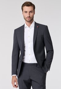 Donkergrijze colbert met een notch lapel, gecombineerd met een witte dress shirt met gouden knopen. Subtiel rood accentdetail op de lapel.