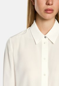 White blouse with a classic collar, featuring subtle texture, button-down front, and a decorative button accent at the collar.