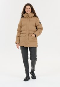 Ung kvinde, der smiler, står iført en tan farvet pufferjakke med høj krave, sorte jeans og sorte kraftige støvler mod en hvid baggrund.