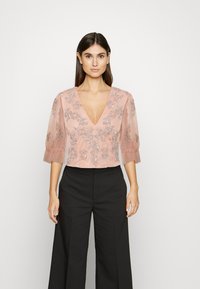 Blusa rosa claro com decote em V profundo, adornada com bordados florais prateados. Mangas curtas semi-transparentes e um design cropped. Usada com calças pretas.