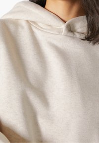 Beige huvtröja gjord av mjukt tyg, med en rundad halsringning och en knappdetalj nära halsen. Slät textur, avslappnad design.