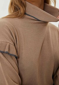 Brauner Rollkragenpullover mit gerippter Textur, der mit Rüsche an den Ärmeln und einem marineblauen Akzent entlang des Kragens und der Ärmelabschlüsse versehen ist.