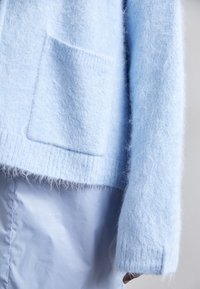 Pull doux bleu clair avec une poche rectangulaire à l'avant et une jupe ou un pantalon en tissu bleu clair assorti visible sous l'ourlet.