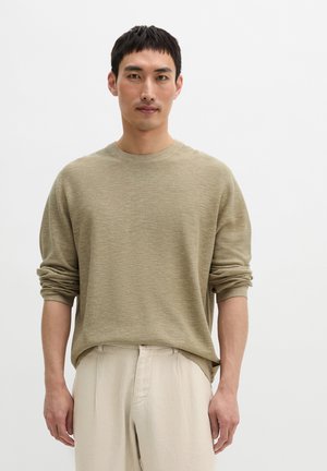 Mężczyzna ubrany w jasnobrązowy sweter z długim rękawem i beżowe spodnie, stojący na jednolitym białym tle.