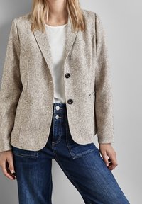 Blazer beige texturé avec un col cranté, doté de deux poches avant et de deux boutons. Assorti à un jean taille haute bleu foncé.