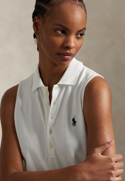 Biała bezrękawnikowa koszulka polo z prążkowaną strukturą, pięcioma guzikami i małym granatowym logo na lewej piersi. Ramię modela skrzyżowane.