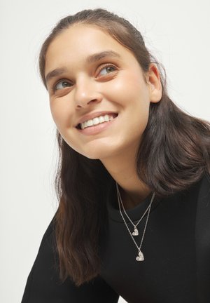 Joven con cabello largo y oscuro sonríe mientras lleva dos collares con colgantes plateados en forma de corazón sobre una blusa negra, con fondo blanco.