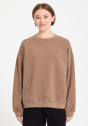 Jeune personne portant un sweat-shirt marron ample et un pantalon noir, debout et souriant devant un fond blanc uni.