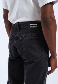 Jeans de mezclilla negros con un corte clásico, con una etiqueta de cuero en la cintura que lleva el nombre de la marca "DRDENIM" y bolsillos traseros.