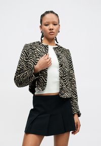 Chaqueta con estampado de cebra en tela suave, diseño corto con escote redondo y dos botones. Se lleva sobre una blusa blanca y combinada con una falda negra.