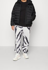 Black padded jacket with a hood, paired with loose white sweatpants featuring a black tie-dye pattern, and light-colored athletic shoes.