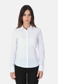 Camicia bianca a maniche lunghe con colletto a punta, vestibilità su misura e texture liscia, abbinata a pantaloni neri.