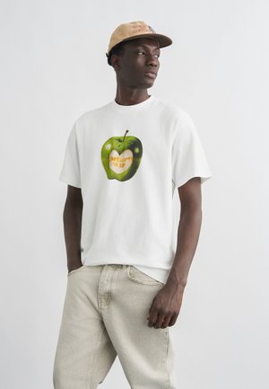 Joven con gorra beige, camiseta blanca con gráfico de manzana verde que contiene el texto "Carhartt WIP", y jeans de color claro sobre un fondo liso.