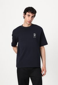 T-shirt en coton bleu marine à manches courtes, col rond et petite illustration blanche sur la poitrine gauche. Présente une coupe décontractée.