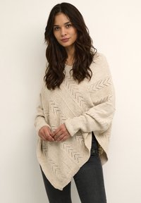 Beige knitted poncho with a chevron pattern, featuring open-knit accents and side slits. Long sleeves and a relaxed fit.