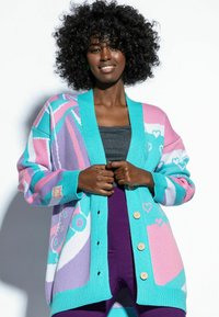 Kardigan w wielokolorowym splocie, z geometrycznymi wzorami, turkusowym ścięciem z prążków i drewnianymi guzikami. Nosi go z szarym topem i fioletowymi legginsami.
