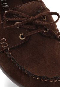 Mocasines de ante marrón con punta redonda, detalle de cordones, ojales metálicos y costuras en contraste a lo largo del borde. Textura suave, suela plana.