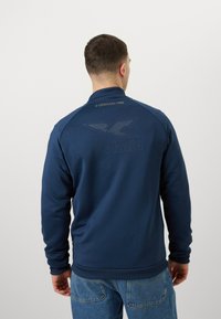 Sweat-shirt bleu marine avec un col et des poignets côtelés, arborant un logo en relief et un texte doré au dos.