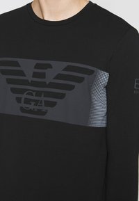 Camisa de manga larga negra con un gran emblema de águila gris, un panel texturizado en el pecho y un patrón gráfico sutil en la manga.