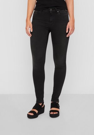 Kobieta w czarnych dopasowanych dżinsach i czarnych sandałach na koturnach stojąca na jednolitym jasnym tle.