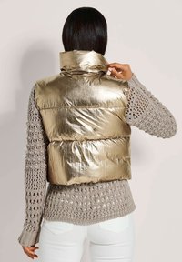 Goldener Puffer-Weste mit hohem Kragen und glänzender Textur, getragen über einem grauen Strickpullover. Verfügt über gepolsterte Segmente und eine enganliegende Passform.