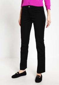 Černé strečové kalhoty s úzkým střihem, které mají přední zapínání na knoflík a kapsy, v kombinaci s černými polobotkami. Hladká textura látky.