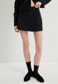 Minifalda negra con un tejido texturizado, que presenta dos botones en la parte delantera y bolsillos laterales. Combinada con calcetas negras y zapatos.