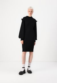Black ribbed knit dress with a high neckline, long puff sleeves, and a fitted silhouette, paired with chunky black shoes and white socks.