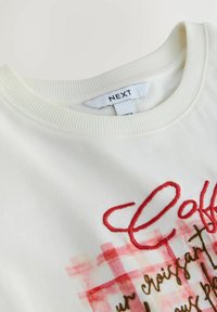 Kremfarvet bomulds-sweatshirt med ribbet krave, med rødt broderet tekst og grafisk design med ternet mønster i bløde farver.