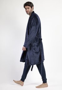 Hombre caminando descalzo, vistiendo una bata suave de color azul marino con cinturón y bolsillos, combinada con pantalones jogger oscuros, frente a un fondo blanco liso.